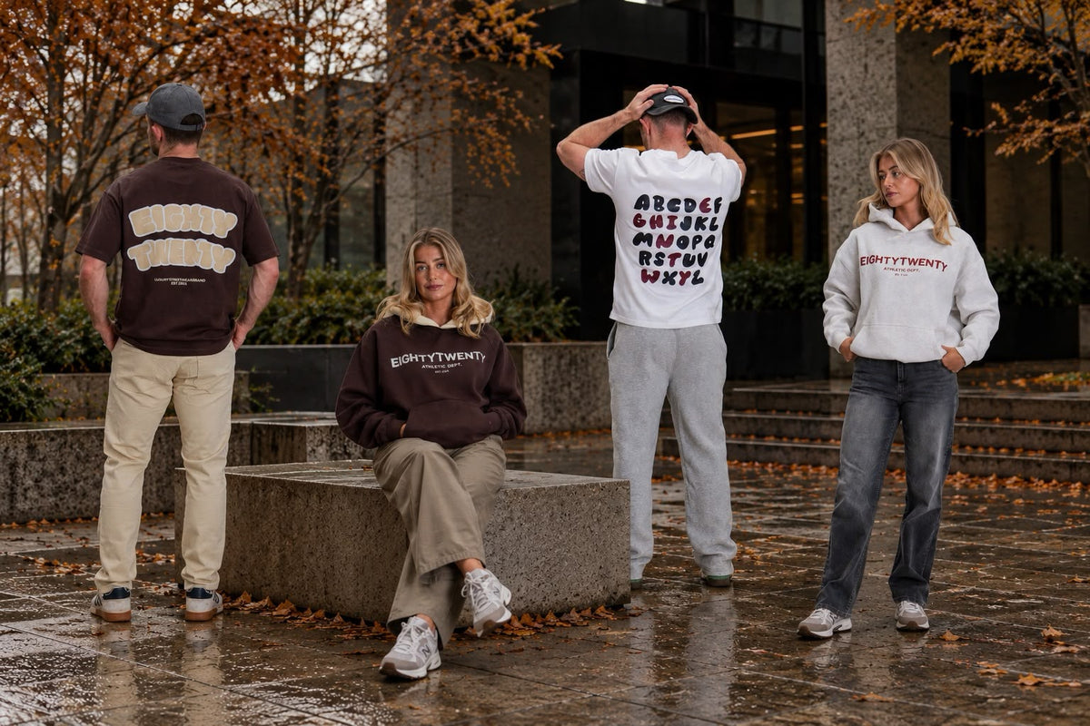 Four people modeling apparel outdoors on a wet stone plaza with autumn trees in the background. One person wears a dark brown sweatshirt with a white graphic, another wears a white sweatshirt with black text, a third wears a dark brown T-shirt with a white graphic, and a fourth wears a white sweatshirt with a red graphic.